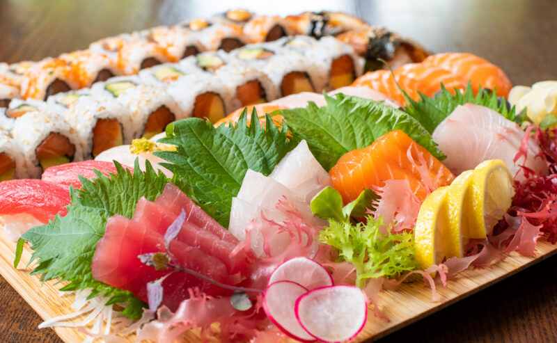Hurley House - A wooden platter featuring assorted sashimi slices including tuna, salmon, and yellowtail, garnished with lemon, greens, and radishes, with sushi rolls in the background.