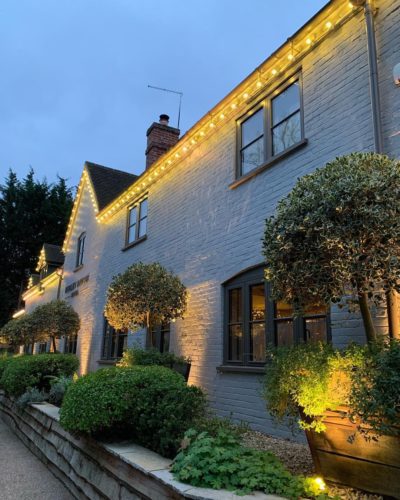 Hurley House - A charming brick building with several windows is adorned with small string lights along the roofline. Potted topiary trees and lush green bushes line the pathway in front, creating a warm, inviting atmosphere under a dusky sky.