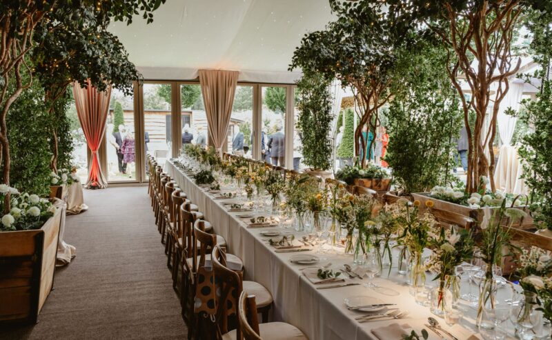 Hurley House - A long, elegant banquet table is set for a formal event, decorated with flowers, greenery, and candles under a canopy with string lights. Large windows and trees create a lush, garden-like atmosphere. People gather outside.
