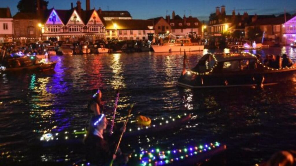 Hurley House - Boats decorated with colorful lights float on a river at night, reflecting vibrant colors on the water. People in kayaks also adorned with lights paddle in the foreground, while illuminated buildings line the riverbank.