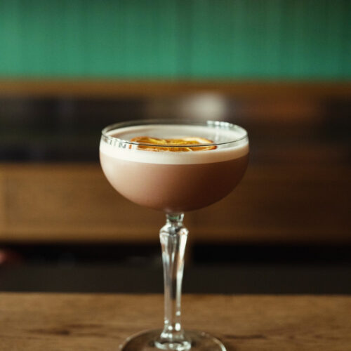 Hurley House - A coupe glass filled with a creamy, light brown cocktail topped with a dried citrus slice, set on a wooden surface with a blurred green and brown background.