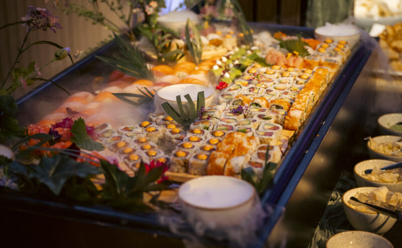 Hurley House - A large platter of assorted sushi rolls and nigiri, garnished with green leaves and flowers, is displayed on a bed of ice at a buffet in a luxury wedding venue in Berkshire, with several small bowls of dipping sauces nearby.