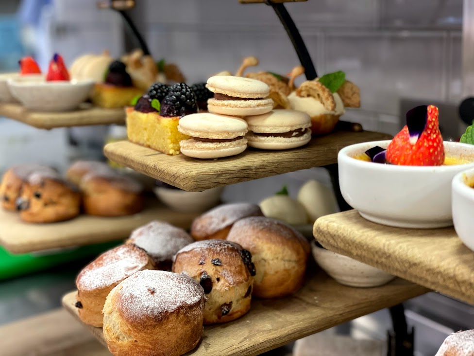 Hurley House - A variety of pastries, including scones, macarons, cakes, and fruit-topped desserts, are artfully arranged on wooden display stands in a bakery or café setting.