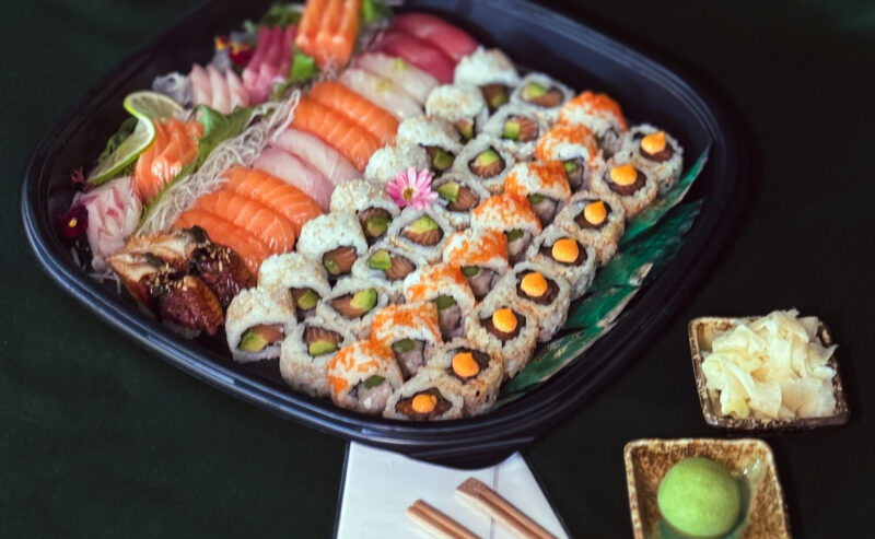 Hurley House - A platter of assorted sushi and sashimi with rolls, nigiri, and garnishes on a green tablecloth. Chopsticks, wasabi, pickled ginger, and two menus are set beside the tray. A small floral arrangement is in the background.