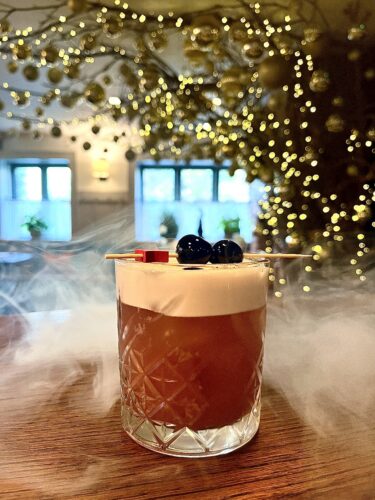 Hurley House - A cocktail with foam in a patterned glass, garnished with two dark cherries on a stick, sits on a wooden table. Mist surrounds the glass, with blurred golden lights and windows in the festive background.