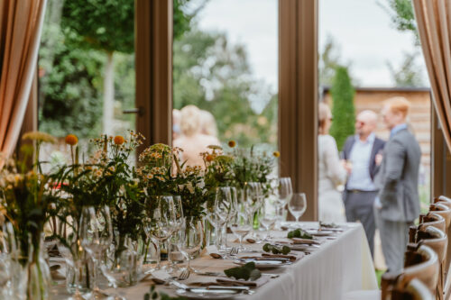 Hurley House - A long dining table set with glassware, cutlery, and floral centerpieces sits indoors near open doors, with a blurred group of people talking outside in a garden setting.