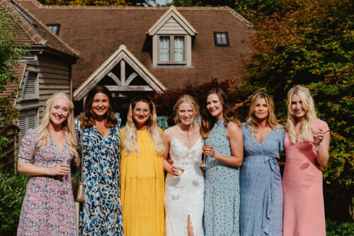 Hurley House - Seven women stand close together, smiling outdoors in front of a house. They wear colorful dresses, and one in the center is in white bridal attire, suggesting a wedding celebration. Sunlight and greenery surround them.