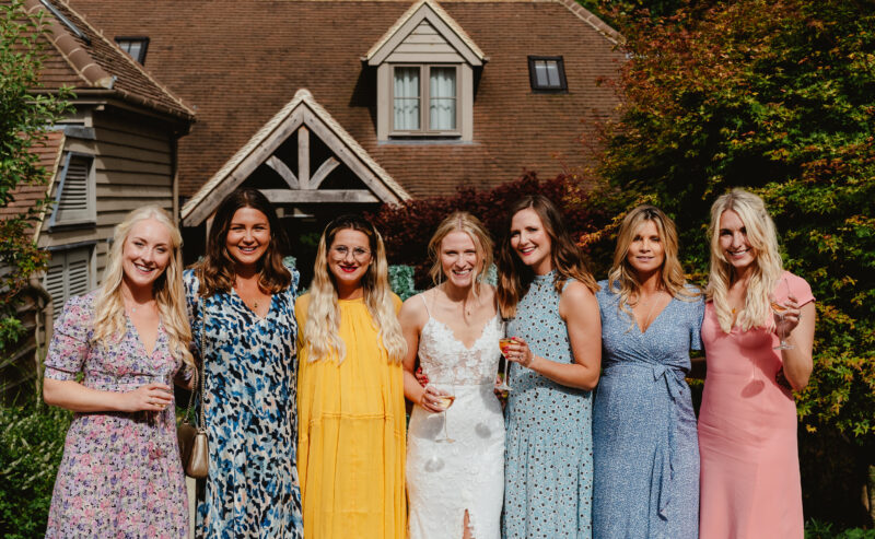 Hurley House - Seven women smiling in colorful dresses stand in front of a stunning luxury wedding venue in Berkshire, featuring a triangular roof and gabled windows. One woman, dressed in white, appears to be the bride. They are outdoors, surrounded by lush greenery and trees.