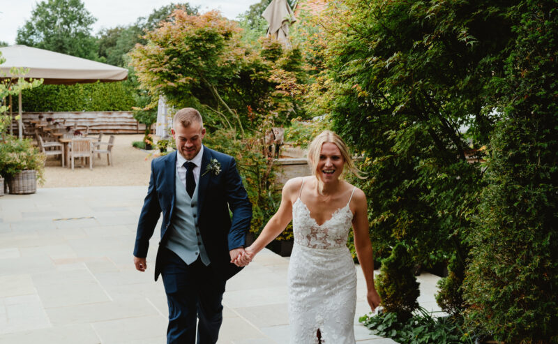 Hurley House - A joyful couple walks hand in hand on a stone path outdoors at a luxury wedding venue in Berkshire. The man is wearing a dark suit, and the woman is in a white wedding dress. They’re surrounded by lush greenery, and patio furniture is visible in the background.