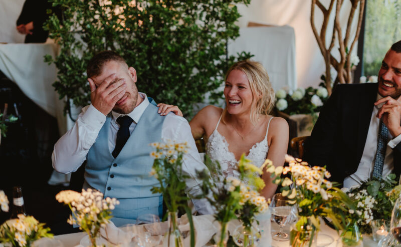 Hurley House - A couple sits at a table adorned with flowers at a luxury wedding venue in Berkshire. The man, in a light blue vest and tie, hides his face with his hand as the woman, wearing a white lace dress, laughs and rests her hand on his shoulder. Trees create a blurred background.