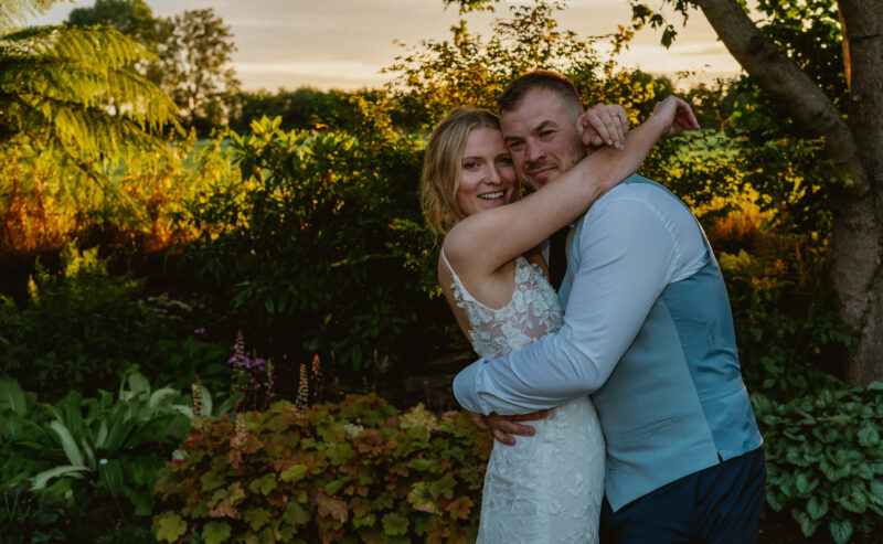Hurley House - A couple embraces in a luxury wedding venue in Berkshires garden during sunset. The woman, in a white lace dress, smiles while the man, in a blue vest, hugs her tightly. They are surrounded by lush greenery and colorful plants, with a warm sky in the background.