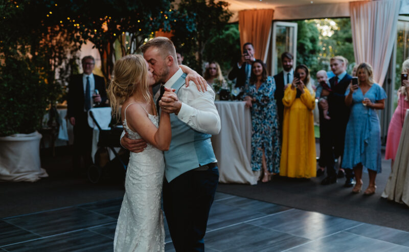 Hurley House - At a luxury wedding venue in Berkshire, a bride and groom share their first dance. Guests stand around watching, some taking photos. The elegantly decorated space, adorned with trees and draped curtains, creates a festive atmosphere perfect for this unforgettable moment.