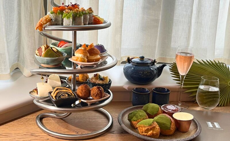 Hurley House - A modern three-tiered stand holds assorted sushi, pastries, and savory bites. Nearby are matcha scones with jam and cream, a teapot, a glass of pink sparkling drink, and a glass of water on a wooden table by a window.