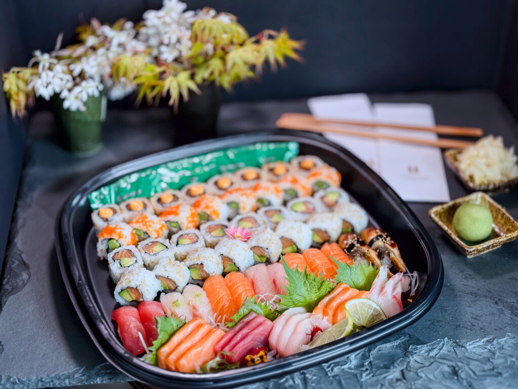 Hurley House - A platter of assorted sushi and sashimi, including salmon, tuna, and various sushi rolls, is displayed on a dark surface with garnishes. Chopsticks, pickled ginger, and wasabi are placed nearby, with flowers in the background.