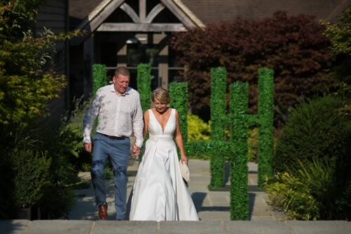 Hurley House - A couple walks hand in hand through a garden at a luxury wedding venue in Berkshire. The woman, in a white wedding dress, and the man, dressed in a patterned shirt and blue pants, meander amid lush greenery and manicured bushes, with an elegant building behind them.