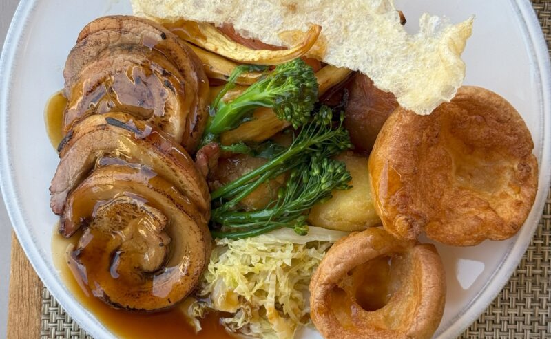 Hurley House - A plate of roast pork slices with gravy, Yorkshire puddings, broccoli, cabbage, roasted potatoes, and a crisp flatbread, served on a white plate with a glass of water nearby.
