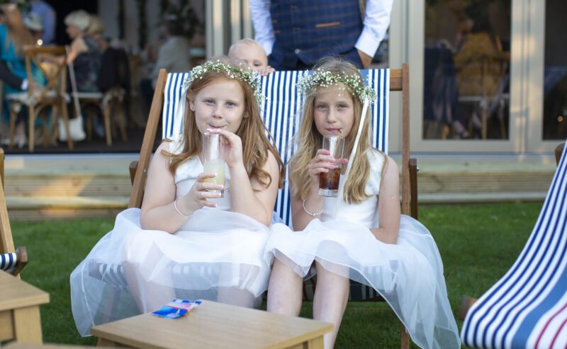Hurley House - Two young girls in white dresses and floral headbands sit on striped deck chairs, sipping from bottles with straws. A baby peeks from the background, while a man in a vest stands nearby. This charming scene unfolds at a luxury wedding venue in Berkshire, surrounded by tables and guests outdoors.