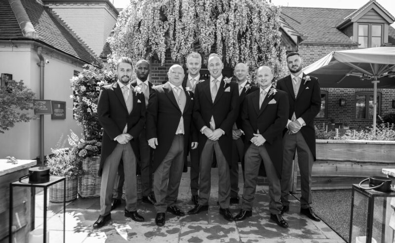 Hurley House - A group of eight men in formal suits stand together outdoors, posing for a photo at a luxury wedding venue in Berkshire. They are in front of a lush, white flowering tree on a sunny day, with brick buildings and a patio area elegantly framing the backdrop.