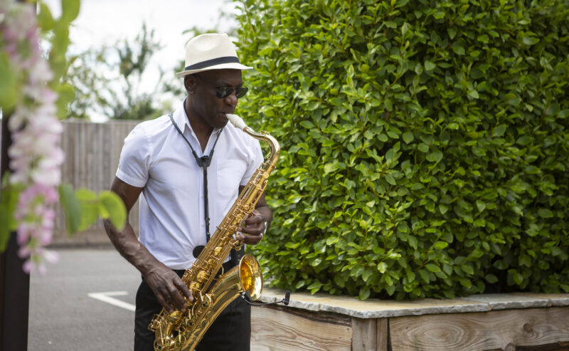 Hurley House - A man in a white shirt and hat plays a saxophone outdoors near a lush green hedge, as if setting the serene tone for a luxury wedding venue in Berkshire.