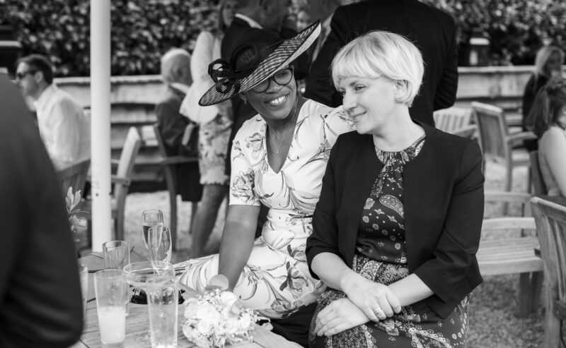 Hurley House - Two women sit and smile at each other outdoors during a social gathering at a luxury wedding venue in Berkshire. One wears a floral dress and hat, the other a patterned dress and blazer. Glasses of drinks are on the table before them, with people and foliage in the black-and-white background.