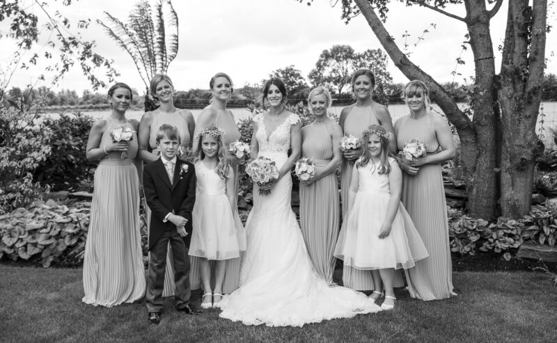 Hurley House - A bride in a wedding dress stands with her bridesmaids and three children in a garden at a luxury wedding venue in Berkshire. The bridesmaids wear matching dresses, while the children are dressed formally: two girls in dresses and a boy in a suit, with lush trees and foliage as their backdrop.