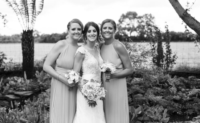 Hurley House - Three women smile while standing together in a garden at a luxury wedding venue in Berkshire. The woman in the center wears a wedding dress, while the other two wear matching bridesmaid dresses and hold small bouquets. The background features greenery and a slightly cloudy sky.