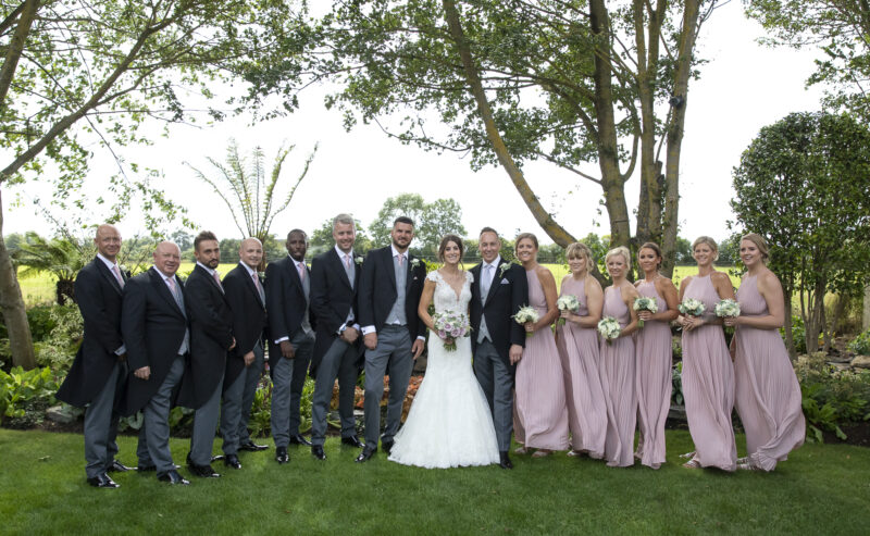 Hurley House - A wedding party poses outdoors at a luxury wedding venue in Berkshire, under lush trees. The bride, in a white gown, stands with the groom and groomsmen in gray suits on one side, while bridesmaids in lilac dresses are on the other. They all smile with greenery as their backdrop.