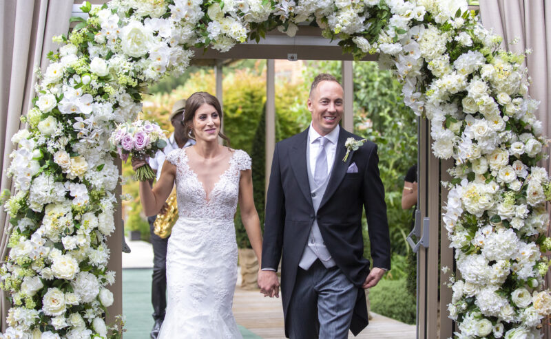 Hurley House - A couple strolls hand in hand under a floral archway adorned with white flowers at a luxury wedding venue in Berkshire, holding a bouquet of pink and purple blooms. The bride dazzles in a lace gown, while the groom looks sharp in a black suit with a white tie and boutonniere.
