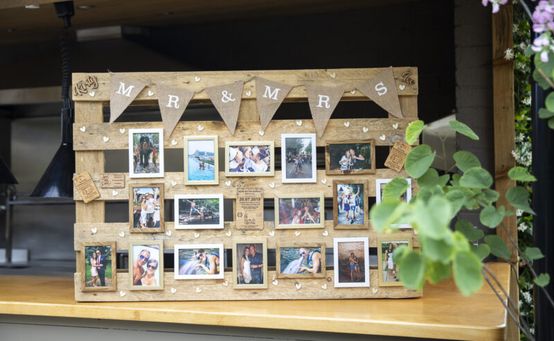 Hurley House - A wooden photo display board with Mr & Mrs at the top showcases a couples cherished moments, adorned with wooden hearts and burlap flags. Set on a kitchen counter with nearby plants, its a charming nod to their Luxury Wedding venue in Berkshire.