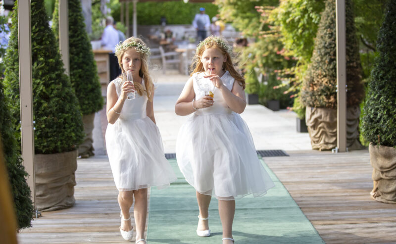 Hurley House - Two young girls, adorned in white dresses and flower crowns, stroll side by side on a wooden pathway at a luxury wedding venue in Berkshire. They sip drinks through straws, surrounded by potted trees and lush greenery.