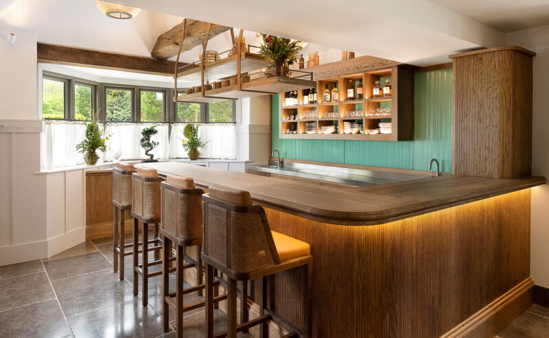 Hurley House - A cozy, modern kitchen reminiscent of an Omakase Sushi Bar—the best in Berkshire—boasts a wooden bar counter flanked by bar stools and open shelves holding jars and plates. The room features a large window with curtains, a green accent wall, and warm lighting.