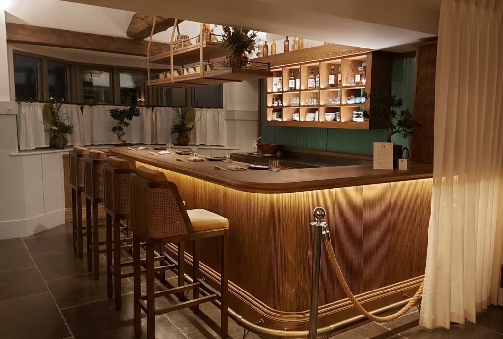 Hurley House - A warmly lit, empty bar with high wooden chairs, a wooden counter, shelves with bottles and glasses, greenery accents, and a rope barrier in front. The atmosphere is cozy and inviting.