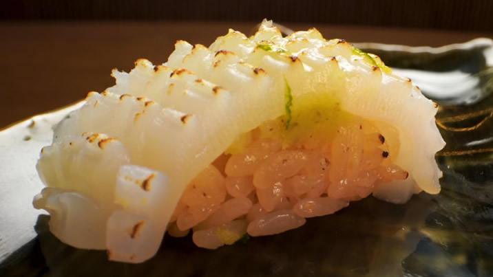 Hurley House - A close-up of a piece of sushi featuring seared, scored white fish on top of vinegared rice, garnished with a hint of green zest, served on a dark ceramic plate.
