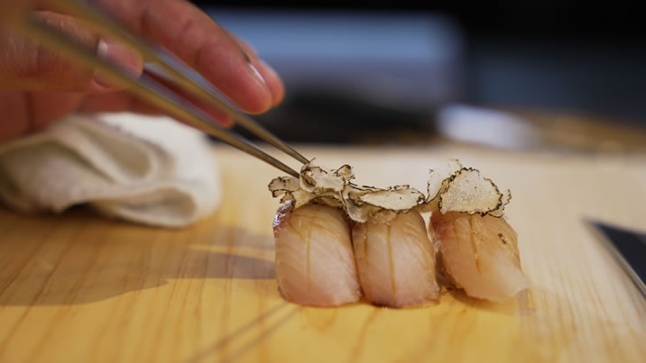 Hurley House - A hand uses metal chopsticks to place thin truffle slices on top of three pieces of sushi arranged on a wooden surface.