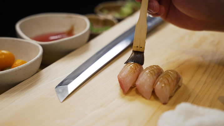 Hurley House - A hand uses a brush to apply sauce to three pieces of nigiri sushi on a wooden board, with a knife and bowls of ingredients in the background.
