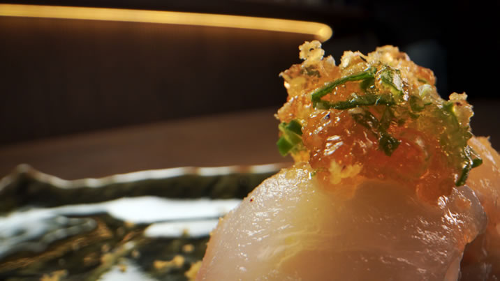 Hurley House - A close-up of a sushi piece topped with finely chopped green herbs and crispy golden garnish on a rustic plate, with a blurred background.