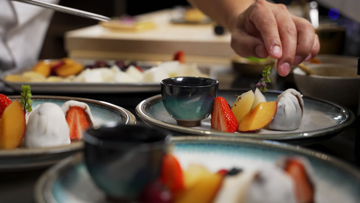 Hurley House - A chef carefully garnishes plates of assorted desserts, featuring fresh strawberries, sliced peaches, and small cups, on a blue-rimmed plate. The background shows more dishes and a blurred kitchen setting.
