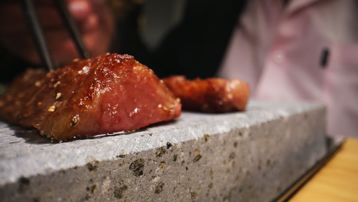 Hurley House - A close-up of someone using chopsticks to pick up a piece of seared meat from a hot stone slab, with another piece of meat and a person in a pink shirt visible in the background.