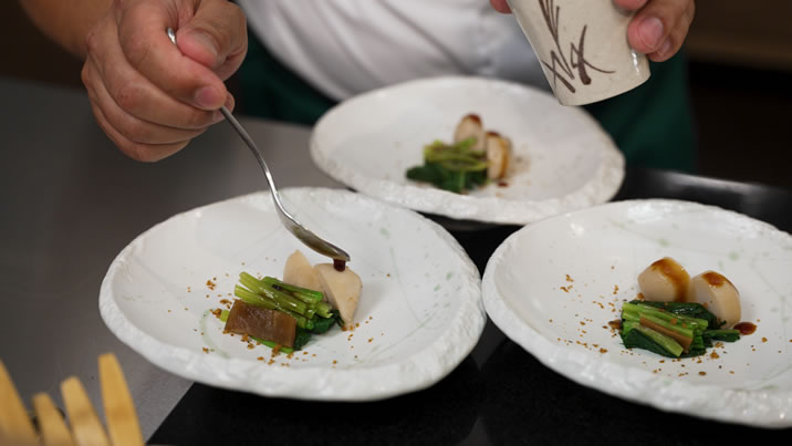 Hurley House - A chef carefully plates a gourmet dish, drizzling sauce over vegetables and scallops arranged artfully on three white plates.