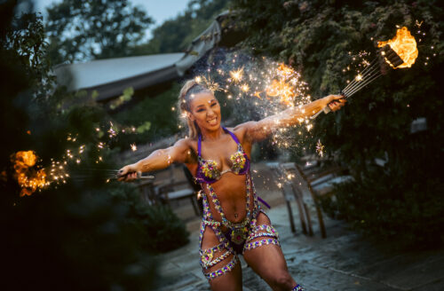 Hurley House - A woman in a vibrant, sequined costume performs outdoors at dusk, holding two fire fans with sparkling flames, smiling energetically amidst lush greenery.