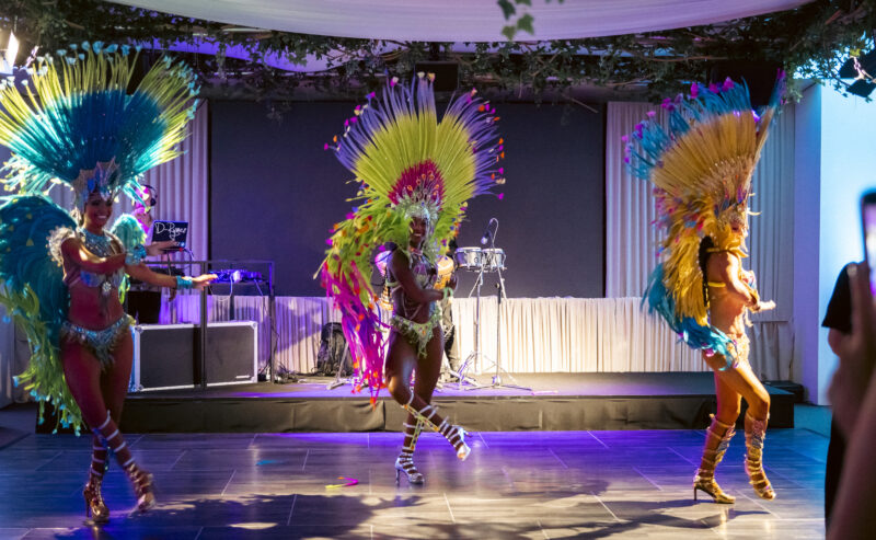 Hurley House - Three dancers in colorful feathered costumes perform energetically on a stage with vibrant lighting. The background has musical instruments and decorations with greenery above. An audience member is visible in the foreground.