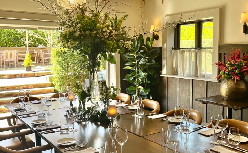 Hurley House - A sunlit dining room with a large wooden table set for a meal, leather chairs, glassware, cutlery, and napkins. Floral centerpieces and plants decorate the room, with round ceiling lights overhead and large windows letting in light.