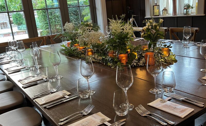 Hurley House - A dining table elegantly set for a meal, with glasses, napkins, and cutlery. The centerpiece features greenery and small orange candles. Large windows with views of a garden provide natural light in the room.