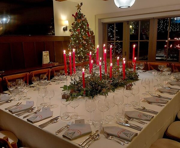 Hurley House - A large square dining table is elegantly set for a holiday meal, featuring red candles, greenery centerpieces, and festive napkins. A decorated Christmas tree and glowing lights are visible in the background.