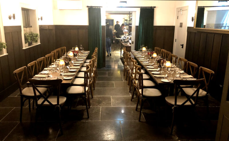 Hurley House - A long dining table set for a formal meal with candles, glassware, and plates, surrounded by wooden chairs in a warmly lit room with dark floors and green curtains. Two people stand near the back.