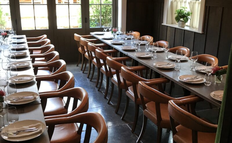 Hurley House - A dining room with two long tables set with plates, glasses, and cutlery. There are leather chairs around the tables, large windows, and modern pendant lights hanging from the ceiling.
