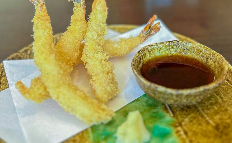 Hurley House - Four pieces of crispy tempura shrimp are served on white paper with a small bowl of dipping sauce and a dollop of wasabi on a rustic ceramic plate.