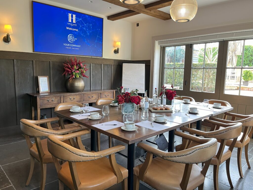 Hurley House - A conference room with a wooden table set for a meeting, featuring notepads, cups, bottled water, and red flower arrangements. A large welcome screen and sunny windows are visible in the background.
