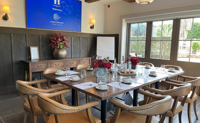 Hurley House - A conference room with a wooden table set for a meeting, featuring notepads, cups, bottled water, and red flower arrangements. A large welcome screen and sunny windows are visible in the background.