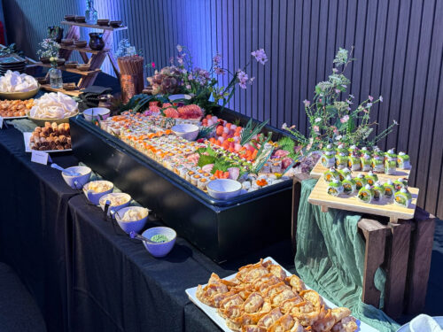 Hurley House - A buffet table displays a large variety of sushi rolls, sashimi, dumplings, and sauces, surrounded by decorative flowers and elegant tableware, set against a wooden wall with blue lighting.